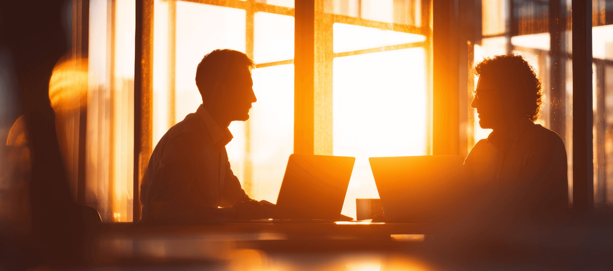 Zwei Männer sitzen im Gegenlicht der untergehenden Sonne am Schreibtisch, Laptops sind geöffnet, es findet ein Gespräch statt